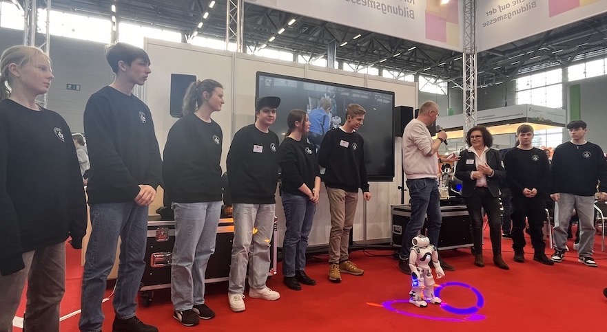 Schülerinnen und Schüler mit Technik-AG-Pullis sowie Lehrerin auf dem Messestand, davor ein Neo-Roboter