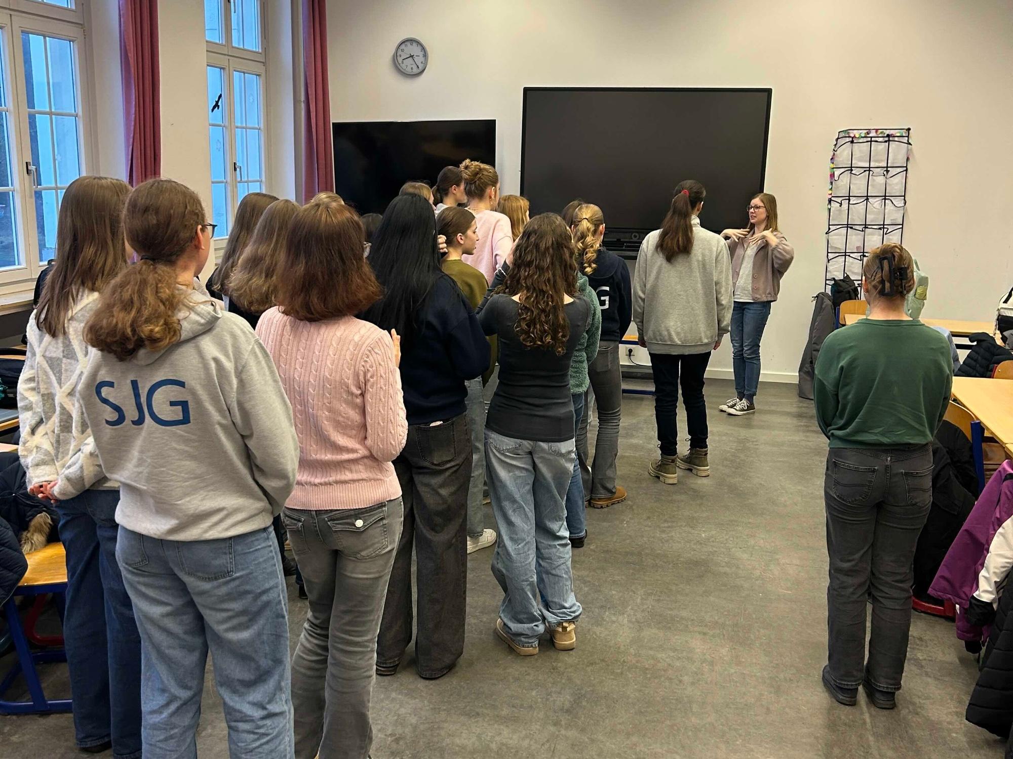 Mädchen stehen in der Klasse und blicken zu einer Frau, die eine Bewegung anzeigt