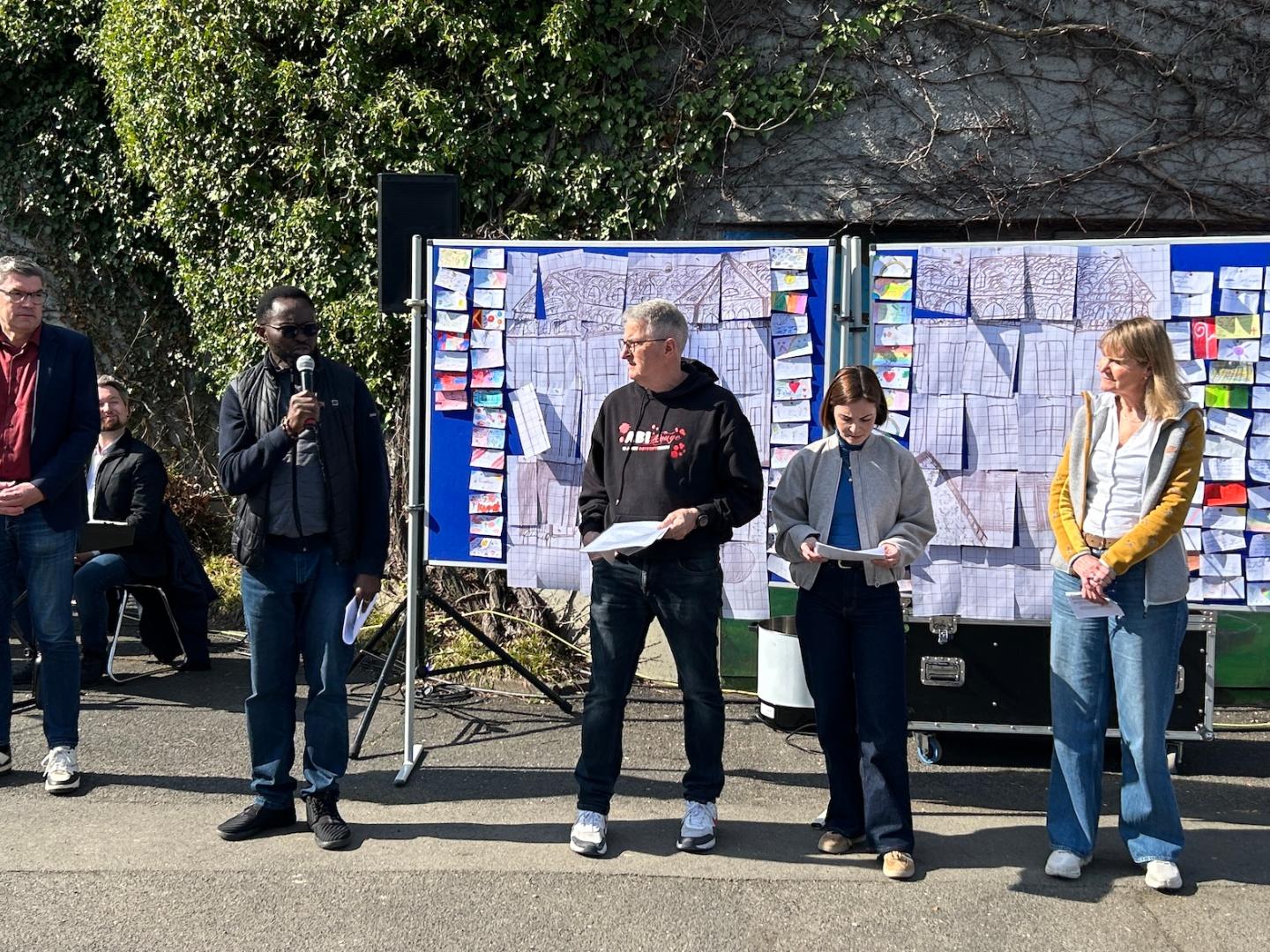 fünf Erwachsene stehen vor einer Stellwand, ein Priester spricht