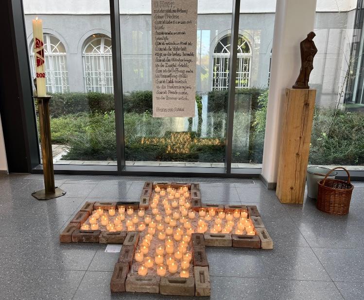 Gedenkkreuz im Foyer Kreuzumriss aus Steinen, darin Sand, darüber ein Gebetstext: Friedensgebet des Franz von Assisi