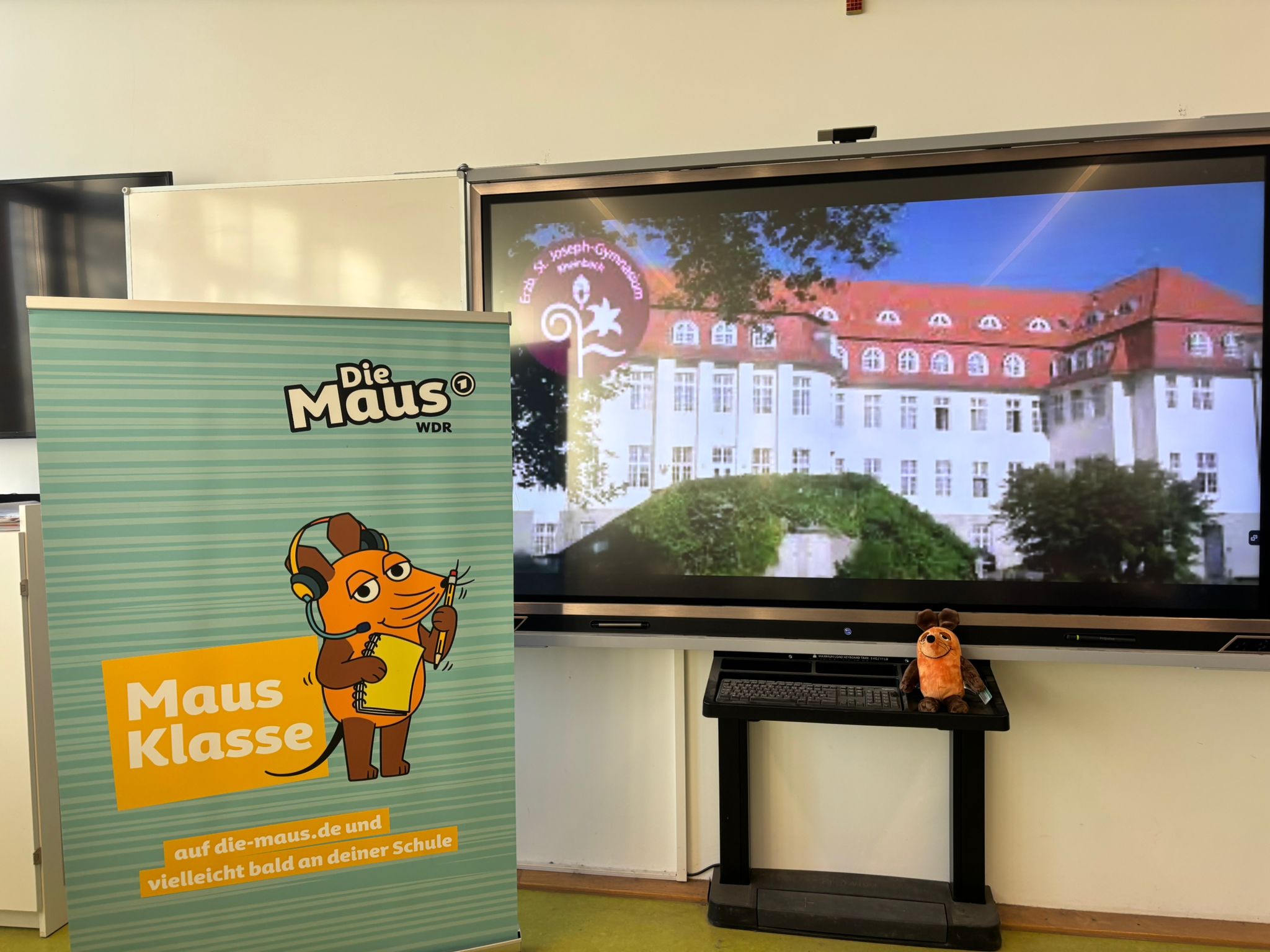 Plakat 'MausKlasse' vor digitalem Board mit Schulbild