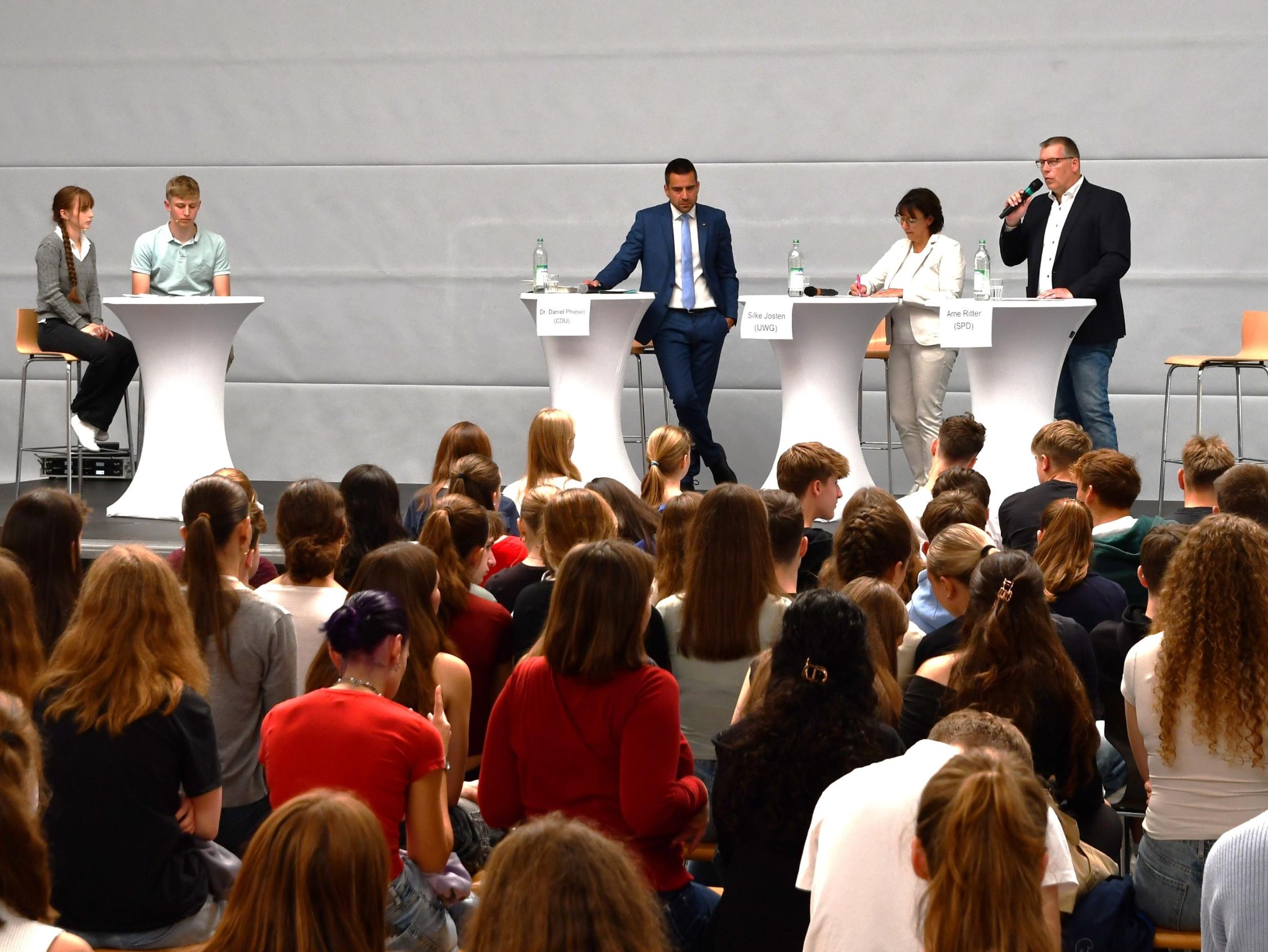 zwei Männer, eine Frau an Stehtischen, einer mit Mikro. Ein weiterer Stehtisch mit einer Schülerin und einem Schüler, Jugendliche