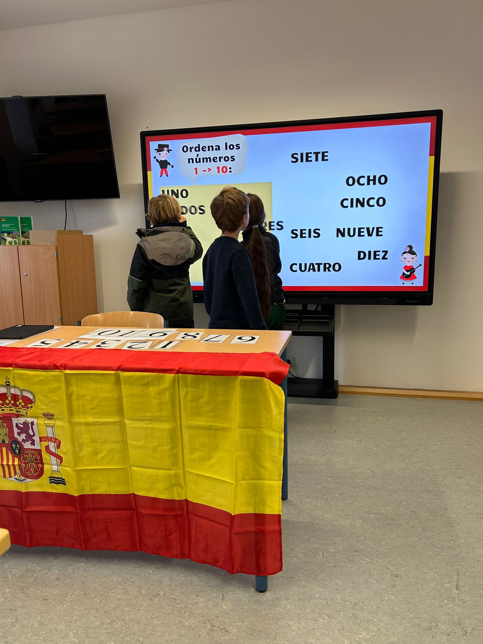 spanische Wörter auf einer digitalen Tafel, davor Kinder und ein Tisch mit einer Spanien-Flagge