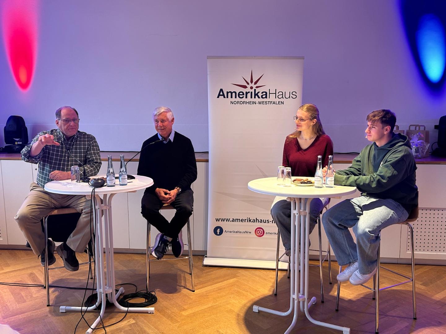 zwei Männer sitzen links auf Barhockern, eine Schülerin, ein Schüler rechts auf Barhockern, dahinter ein Rollup vom Amerika-Haus NRW, rote und blaue Strahler