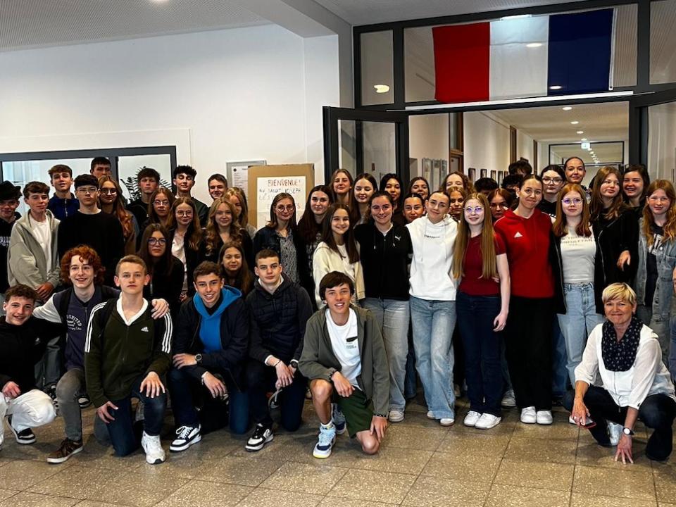 Schülerinnen und Schüler im Schulgebäude unter französischer Flagge, vorne Lehrerinnen und Lehrer