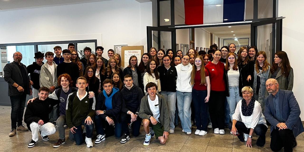 Schülerinnen und Schüler im Schulgebäude unter französischer Flagge, vorne Lehrerinnen und Lehrer